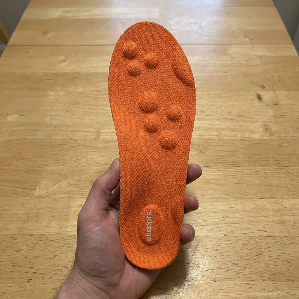 A single orange Stepprs insole held in a hand, close-up on the raised forefoot massage nodes and printed logo