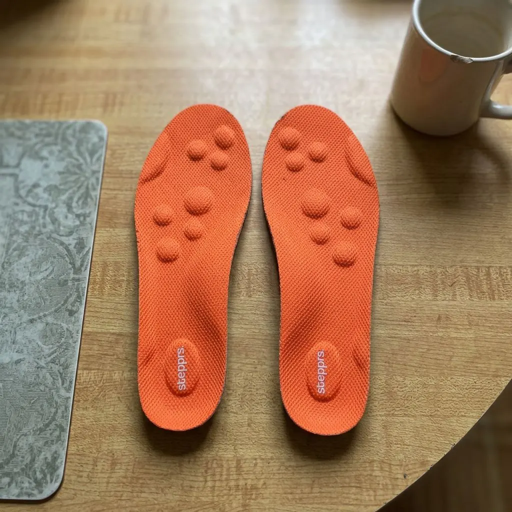 A pair of orange Stepprs insoles laid flat, top-cover side up, showing the raised massage nodes near the forefoot and heel